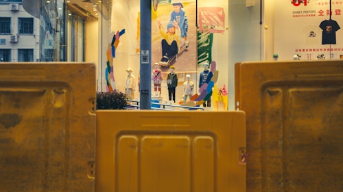 A storefront display featuring mannequins dressed in various clothing items is seen behind yellow barricades. The display includes colorful abstract art and large posters on the window. Outside, there are reflective surfaces and signage visible.