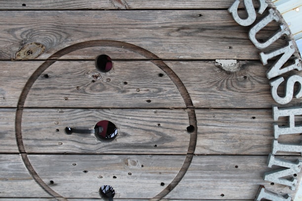 A wooden surface with an abstract, circular pattern cut into it, featuring metallic letters mounted to the right side. The letters spell out a partial word possibly starting with an 'S'. The wood displays natural grain patterns and several holes, giving it a rustic texture.