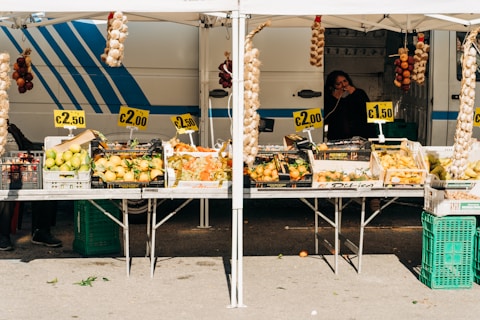 A market stall displays a variety of fresh produce, including lemons, onions, peppers, and grapes, all at different price points. The setup includes signs showing prices like €2.50, €2.00, and €1.50. Hanging above are braided strings of garlic and onions. A person is visible in the background, standing beside a striped van, possibly the vendor.