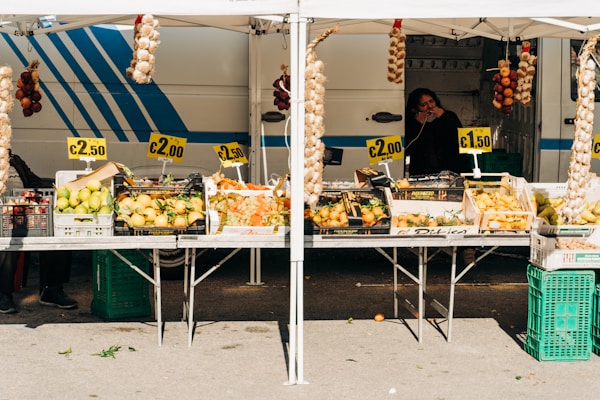 A market stall displays a variety of fresh produce, including lemons, onions, peppers, and grapes, all at different price points. The setup includes signs showing prices like &euro;2.50, &euro;2.00, and &euro;1.50. Hanging above are braided strings of garlic and onions. A person is visible in the background, standing beside a striped van, possibly the vendor.
