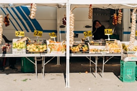 A market stall displays a variety of fresh produce, including lemons, onions, peppers, and grapes, all at different price points. The setup includes signs showing prices like €2.50, €2.00, and €1.50. Hanging above are braided strings of garlic and onions. A person is visible in the background, standing beside a striped van, possibly the vendor.