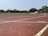 A running track with white lane markings, scattered yellow petals, and a red corner flag. The track curves slightly to the right. Surrounding the track are green grass and a row of trees. Buildings are visible in the background under a clear sky.