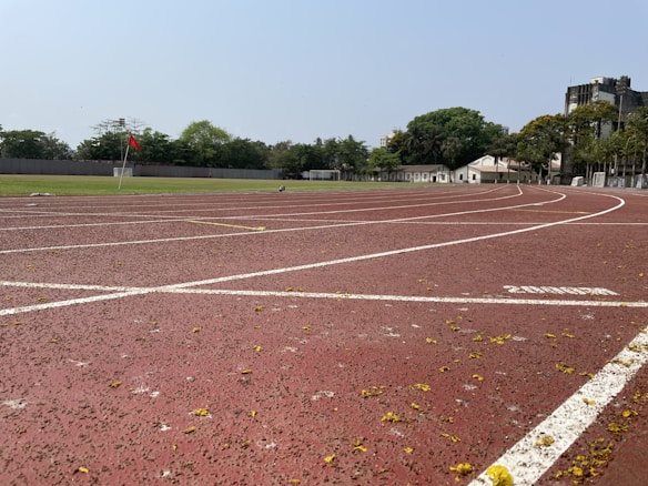 A running track with white lane markings, scattered yellow petals, and a red corner flag. The track curves slightly to the right. Surrounding the track are green grass and a row of trees. Buildings are visible in the background under a clear sky.