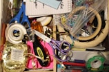 A cluttered drawer with various office supplies including scissors, tape, paper clips, a notepad, and ribbon. There's also a screwdriver and colorful tapes.