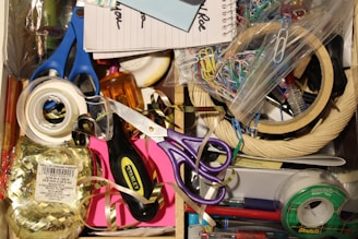 A cluttered drawer with various office supplies including scissors, tape, paper clips, a notepad, and ribbon. There's also a screwdriver and colorful tapes.