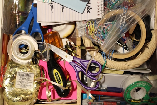 A cluttered drawer with various office supplies including scissors, tape, paper clips, a notepad, and ribbon. There's also a screwdriver and colorful tapes.