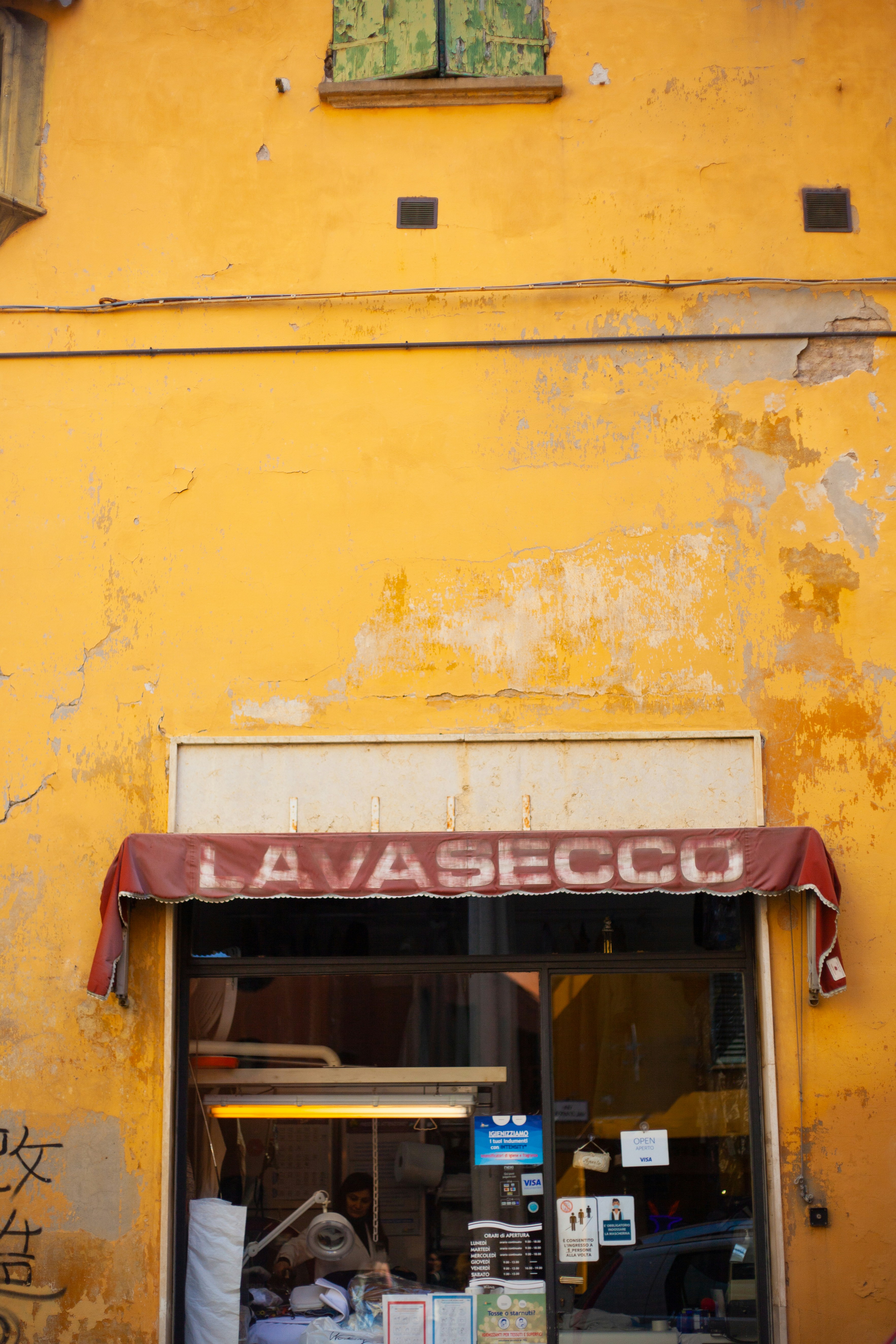 A vibrant yellow, weathered wall with a green, peeling wooden shutter above a storefront. The faded maroon awning displays the word 'LAVASECCO'. Below, the entrance reveals a glimpse into a shop with cleaning equipment visible and informational posters displayed on the glass.