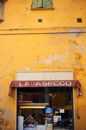 A vibrant yellow, weathered wall with a green, peeling wooden shutter above a storefront. The faded maroon awning displays the word 'LAVASECCO'. Below, the entrance reveals a glimpse into a shop with cleaning equipment visible and informational posters displayed on the glass.