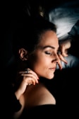 A woman with closed eyes rests peacefully on a pillow, her head slightly tilted and supported by her hand. The lighting is soft, casting gentle shadows across her serene face. Her hair is tied back, and she wears subtle makeup, enhancing her calm demeanor.