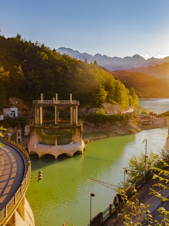A river with vibrant green water flows through a rocky landscape, flanked by dense forests on either side. A small bridge with railings curves over the river, leading to a cylindrical structure by the water's edge. In the background, sunlit mountains with clear skies create a serene and picturesque setting.