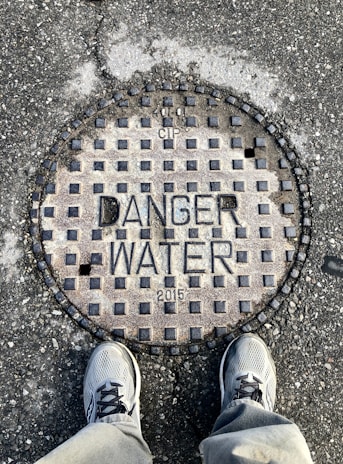A manhole cover displaying the words 'Danger Water 2015' with checkerboard pattern and letters. Two sneakers are visible at the bottom, standing on an asphalt surface.
