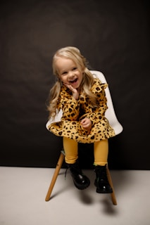 A young child with blonde hair is sitting on a modern chair against a dark background. They are wearing a leopard print dress with mustard yellow leggings and black boots. The child has a cheerful expression and is resting their head on one hand.