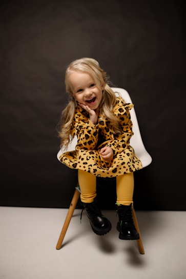 A young child with blonde hair is sitting on a modern chair against a dark background. They are wearing a leopard print dress with mustard yellow leggings and black boots. The child has a cheerful expression and is resting their head on one hand.