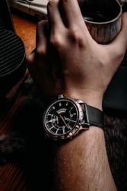 A hand wearing a black leather-strapped watch with a detailed black and silver watch face. The hand is holding a rustic mug, and the background includes a wooden surface and parts of an unidentified object.