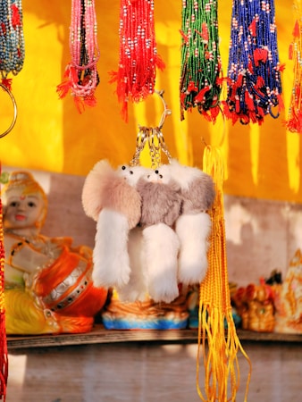 A vibrant display of beaded strings hanging from a stall, predominantly featuring red, green, and blue strands with tassels. A plush, fluffy keychain with a ring is in the center, complemented by decorative items in the background, including a doll-like figure.