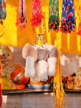A vibrant display of beaded strings hanging from a stall, predominantly featuring red, green, and blue strands with tassels. A plush, fluffy keychain with a ring is in the center, complemented by decorative items in the background, including a doll-like figure.