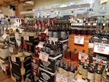 A liquor store display featuring a wide variety of whiskey and bourbon bottles. The shelves are packed with brands like Jack Daniel's, Maker's Mark, and Evan Williams, each with prominent price tags. The background includes decorative neon signs.