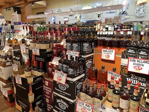 A liquor store display featuring a wide variety of whiskey and bourbon bottles. The shelves are packed with brands like Jack Daniel's, Maker's Mark, and Evan Williams, each with prominent price tags. The background includes decorative neon signs.