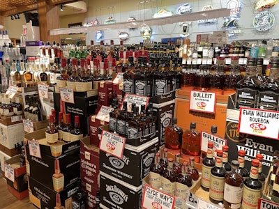 A liquor store display featuring a wide variety of whiskey and bourbon bottles. The shelves are packed with brands like Jack Daniel's, Maker's Mark, and Evan Williams, each with prominent price tags. The background includes decorative neon signs.