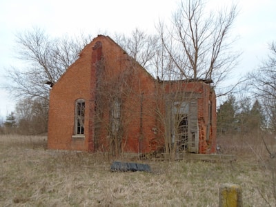 An abandoned brick building stands in a field, with its structure partially deteriorated. The windows are broken, and the roof appears to be collapsing. Leafless trees and overgrown grass surround the building, indicating neglect and decay.
