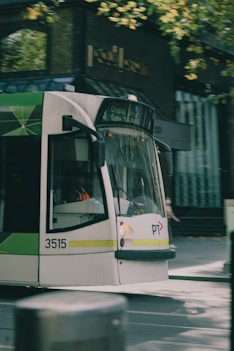 A modern tram in motion travels along a street lined with tall, green trees. The tram's front and side are visible, showcasing its white body with green and gray accents. The background features an urban setting with blurred buildings, suggesting movement and a bustling city environment.