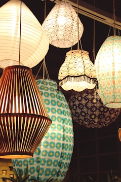 A collection of hanging lamps with various designs and patterns. The lamps include a mix of paper and wooden materials, featuring intricate patterns in colors like white, blue, and brown. The lighting creates a warm and inviting atmosphere, enhancing the decorative patterns on the lamps.