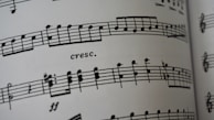 A close-up of a sheet of printed music featuring musical notes and symbols on a staff line. The term 'cresc.' appears, indicating a crescendo. The paper appears to curve slightly, suggesting the image captures a part of an open book.