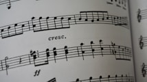 A close-up of a sheet of printed music featuring musical notes and symbols on a staff line. The term 'cresc.' appears, indicating a crescendo. The paper appears to curve slightly, suggesting the image captures a part of an open book.