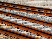 Several parallel railway tracks with a gravel ballast foundation. The tracks are made of steel and show a reddish-brown color, accented by metal fastenings. The scene suggests an industrial or transportation setting, with the sun casting shadows on the rock and metal surfaces.