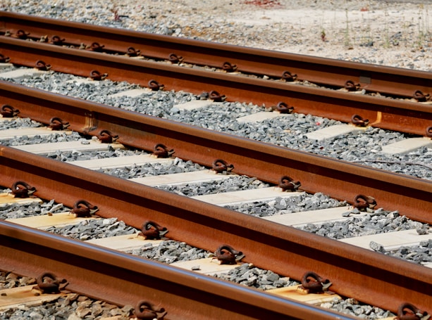 Several parallel railway tracks with a gravel ballast foundation. The tracks are made of steel and show a reddish-brown color, accented by metal fastenings. The scene suggests an industrial or transportation setting, with the sun casting shadows on the rock and metal surfaces.