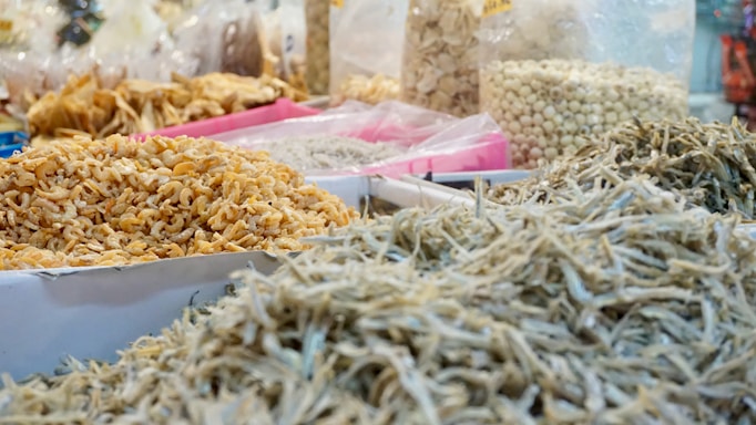 A market stall displays a variety of dried foods including small shrimp, anchovies, and other dried ingredients. Transparent plastic bags filled with different items are visible in the background, along with baskets and trays filled with more dried goods.