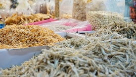 A market stall displays a variety of dried foods including small shrimp, anchovies, and other dried ingredients. Transparent plastic bags filled with different items are visible in the background, along with baskets and trays filled with more dried goods.