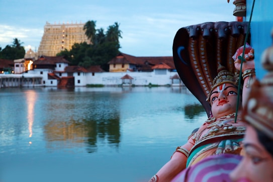 A detailed idol of a deity is positioned on the right side of the image with intricate features and multi-headed hoods of a serpent. In the background, there is a temple with elaborate architecture, surrounded by lush greenery and palm trees. The temple is reflected in a large tranquil body of water, creating a serene atmosphere.