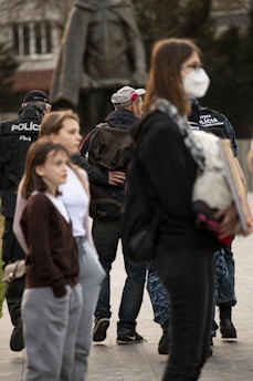 A group of people are gathered outdoors. In the background, officers in black uniforms with the word 'POLICE' are visible, and they appear to be escorting individuals. Two young girls stand in the foreground, with one wearing a brown hoodie and the other a white shirt under a darker jacket. A woman, partially obscured, is wearing a face mask and holding documents or a folder.