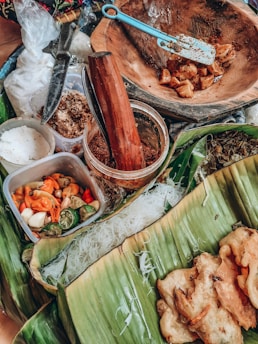 A collection of ingredients and tools for cooking on a green banana leaf. There are sliced chilies, limes, and a container holding a white substance, possibly salt. A wooden mortar with a pestle is present alongside a chopping knife. Some seasoning or spice mix is also visible in a jar. Fried food items are placed on the banana leaf along with strands of rice noodles.