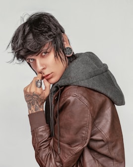 A person with dark hair and a nose piercing wears a large ear gauge and a leather jacket over a gray hoodie. A hand with tattooed art, including a skull design ring, is placed near the chin.