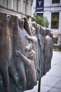 A bronze sculpture depicting a series of human figures emerging from a textured wall, with varied facial expressions and postures, suggesting struggle and perseverance.