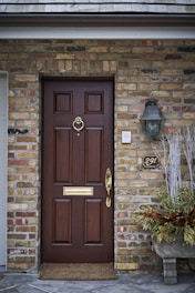 A solid wooden front door features a brass door knocker and handle, set in a brick wall. To the right of the door, a vintage-style wall lantern and a metallic house number 291 are mounted. Below, a rustic doormat lies on a stone patio, and a decorative planter with dry foliage adds an element of greenery.