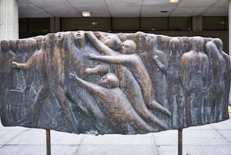 A bronze relief sculpture shows a group of human figures intertwined, some reaching out, while others appear to be merging with a vertical, textured surface. The figures are abstract and expressive, conveying a sense of struggle or connection.