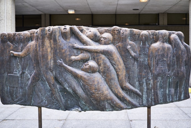 A bronze relief sculpture shows a group of human figures intertwined, some reaching out, while others appear to be merging with a vertical, textured surface. The figures are abstract and expressive, conveying a sense of struggle or connection.