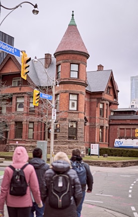 A group of people in winter clothing walk towards a historic red brick building with a rounded tower and pointed roof. The structure features ornate architectural details and arched windows. Street signs indicate the intersection of Harbord Street and Hoskin Avenue, with traffic lights and urban surroundings visible.