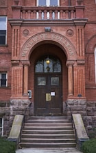 An arched entrance to a brick building with the words 'WYCLIFFE COLLEGE' engraved above dark wooden double doors. Stone steps with metal railings lead up to the entrance. Decorative stonework and columns frame the doorway, and a warm light fixture hangs above the doors.