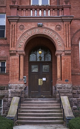 An arched entrance to a brick building with the words 'WYCLIFFE COLLEGE' engraved above dark wooden double doors. Stone steps with metal railings lead up to the entrance. Decorative stonework and columns frame the doorway, and a warm light fixture hangs above the doors.