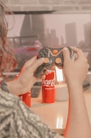 A person is holding a video game controller in front of a monitor displaying a racing game. There are two cans of Coca-Cola and a lamp on a wooden desk. The scene appears to be a cozy gaming setup.