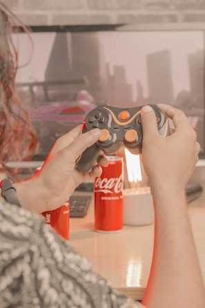 A person is holding a video game controller in front of a monitor displaying a racing game. There are two cans of Coca-Cola and a lamp on a wooden desk. The scene appears to be a cozy gaming setup.