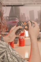 A person is holding a video game controller in front of a monitor displaying a racing game. There are two cans of Coca-Cola and a lamp on a wooden desk. The scene appears to be a cozy gaming setup.
