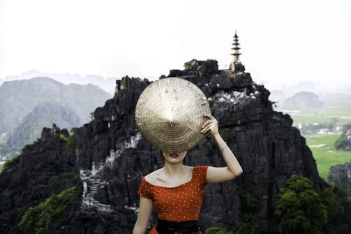 A person wearing a red polka-dot dress is holding a traditional conical hat to cover their face. They stand in front of dramatic rock formations and a pagoda situated atop one of the rocks. The background features lush greenery and a distant view of a landscape with more rock formations under a pale sky.