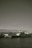 A large industrial ship docked beside a concrete barrier in a harbor setting. The ship features a smokestack and appears to have a camouflaged pattern on its hull. Surrounding the dock are other industrial structures and equipment, partially visible hills, and a partially cloudy sky overhead.