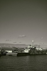 A large industrial ship docked beside a concrete barrier in a harbor setting. The ship features a smokestack and appears to have a camouflaged pattern on its hull. Surrounding the dock are other industrial structures and equipment, partially visible hills, and a partially cloudy sky overhead.