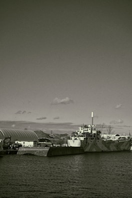 A large industrial ship docked beside a concrete barrier in a harbor setting. The ship features a smokestack and appears to have a camouflaged pattern on its hull. Surrounding the dock are other industrial structures and equipment, partially visible hills, and a partially cloudy sky overhead.